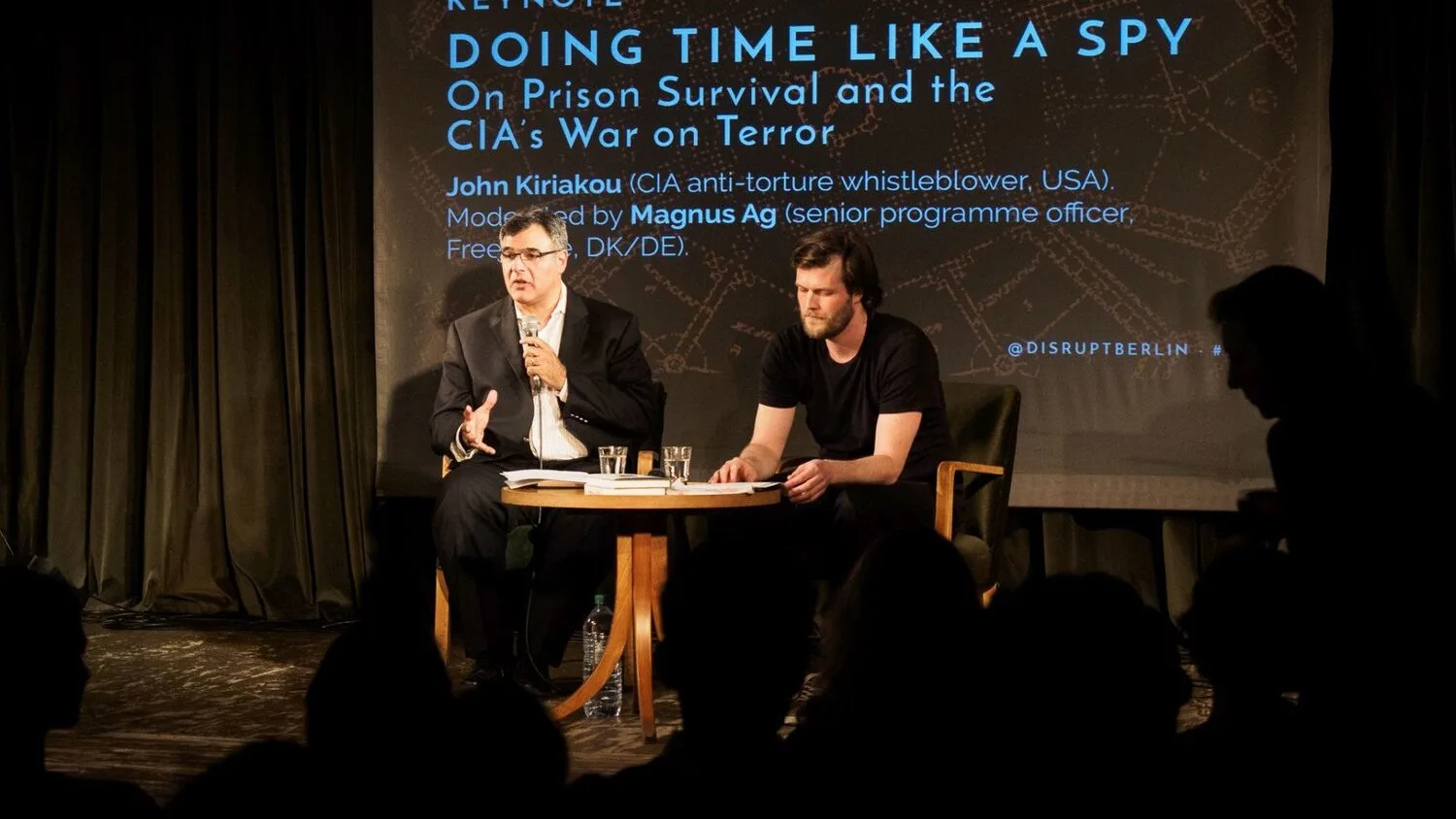 John Kiriakou fala sentado ao lado de um moderador em evento sobre espionagem e tortura da CIA