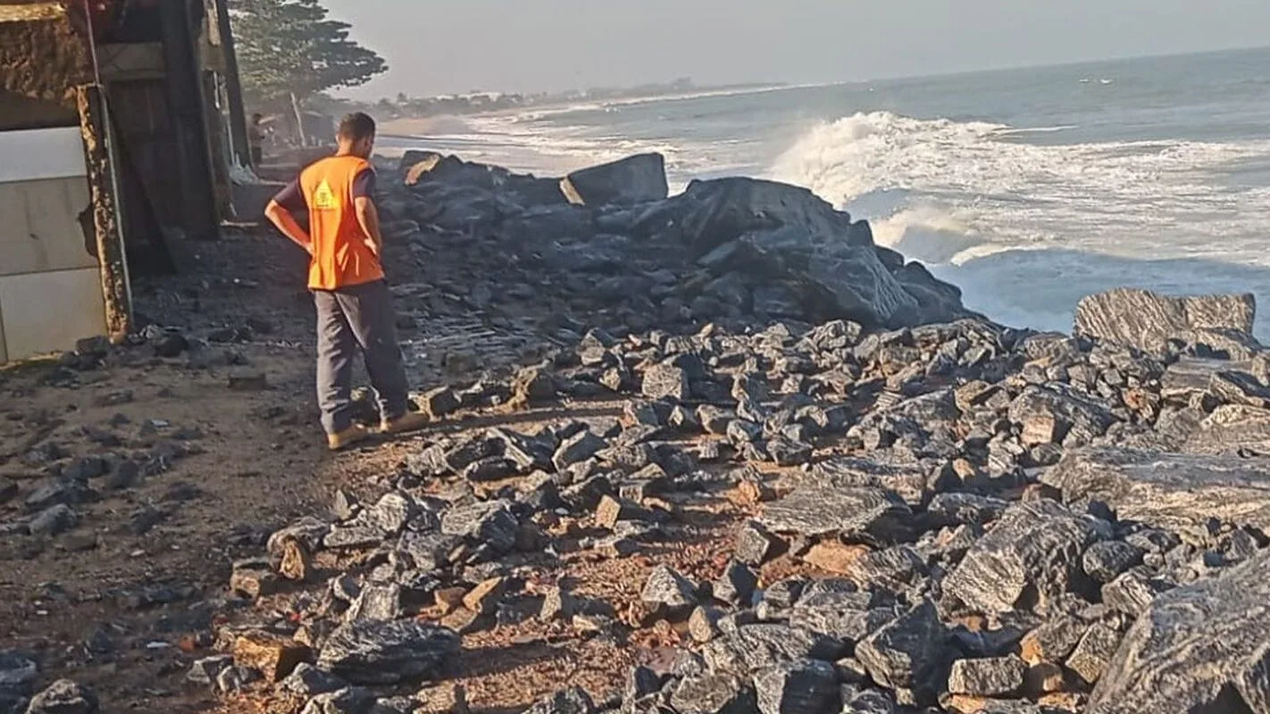 Homem em meio a rochas e destroços na orla, com o mar agitado ao fundo