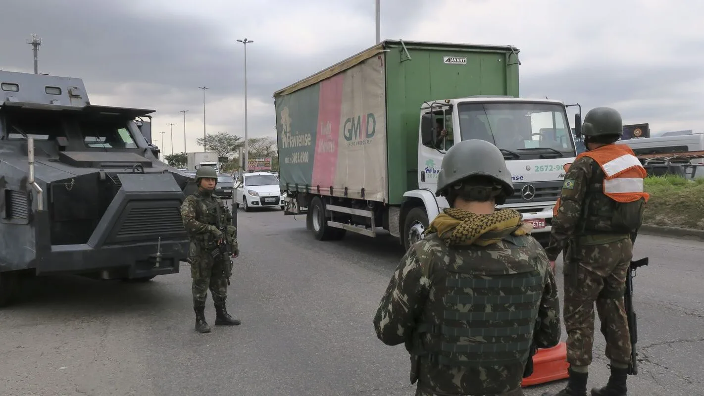 Militares e policiais em operação conjunta contra roubo de cargas no Rio de Janeiro, com blindado e caminhão ao fundo