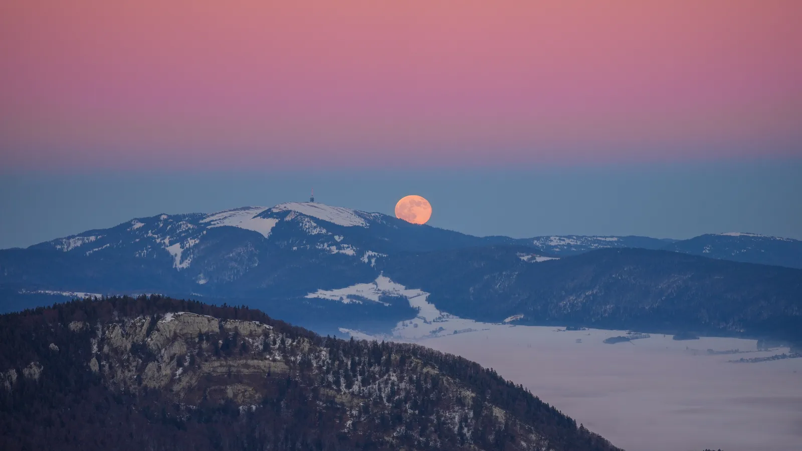 Lua cheia alaranjada surgindo no horizonte ao entardecer, atrás de uma montanha