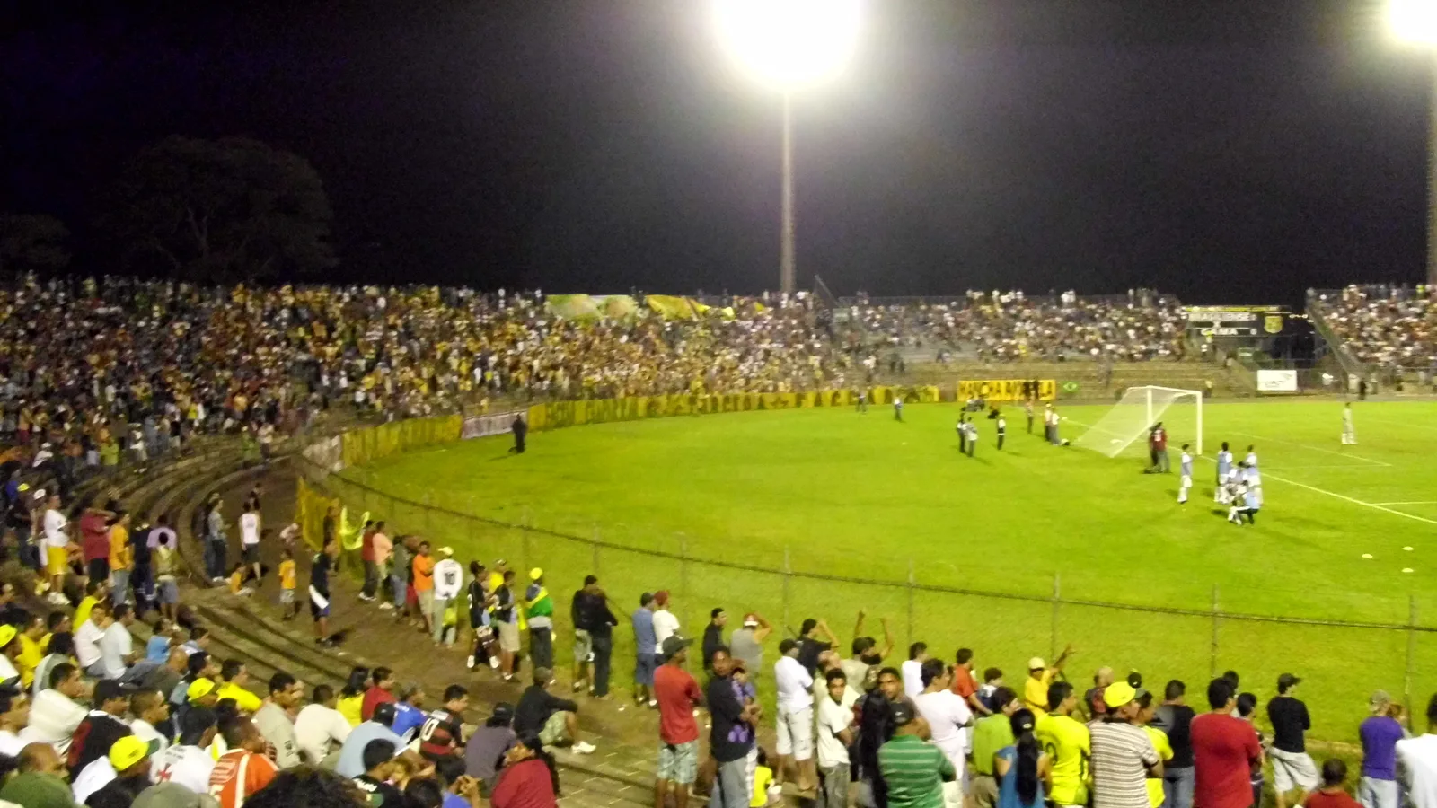Torcida em arquibancada lotada acompanha jogo de futebol à noite sob holofotes