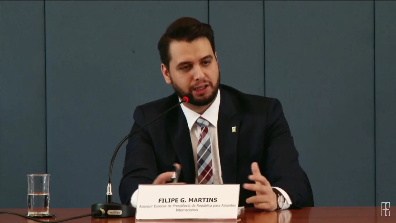Filipe Martins fala ao microfone em painel oficial, identificado por placa com seu nome